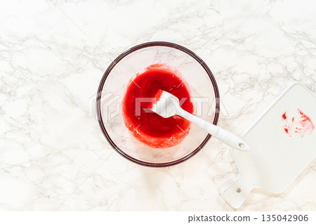 A glass mixing bowl with freshly strained raspberry puree and a spatula resting inside, prepared for use in the Easter Cookie Sandwich with Raspberry Buttercream recipe. Ideal for recipes and 135042906