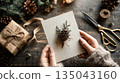 Hands holding a handmade winter greeting card decorated with a pinecone and natural rustic details 135043160