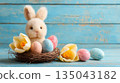 A cute Easter bunny figurine sitting in a nest with colorful eggs on a pastel blue background 135043182