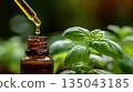 A close up of essential oil dripping from a glass dropper beside fresh basil leaves in natural light 135043185