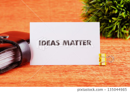 A white card stating IDEAS MATTER sits with a gold clip next to a black book, complemented by a small green plant on an orange surface 135044985