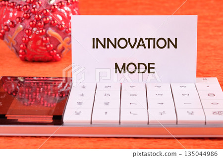 A card stating INNOVATION MODE sits on a white Calculator. Next to it is a decorative red ornament on an orange surface, suggesting fresh perspective 135044986