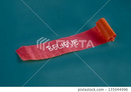A Scarlet paper scroll unrolls across a Teal surface. The word BELIEVE is boldly emblazoned in stark white, a potent symbol of unwavering faith A Scarlet paper scroll unrolls across a Teal surface. The word BELIEVE is boldly emblazoned in stark white, a potent symbol of unwavering faith 135044996