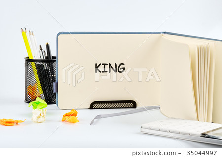 An open notebook displays the word King. Pencils, pens, and crumpled papers scatter around the desk near a book stand. A journal lays near the notebook An open notebook displays the word King. Pencils, pens, and crumpled papers scatter around the desk near a book stand. A journal lays near the notebook 135044997