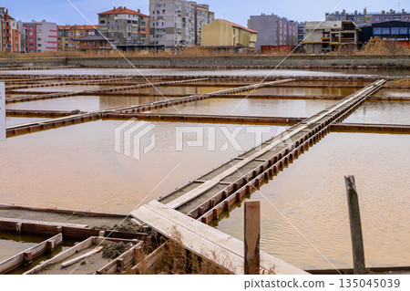 Salt evaporation ponds with wooden frames reflecting sky in Pomorie, Bulgaria, surrounded by residential buildings. Concept of traditional saltwork within modern coastal town 135045039