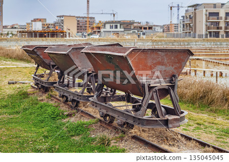 Rusty carts on narrow railway used for salt transport in Pomorie, Bulgaria, stand abandoned near modern construction. Concept of industrial past and changing coastal urbanism Rusty carts on narrow railway used for salt transport in Pomorie, Bulgaria, stand abandoned near modern construction. Concept of industrial past and changing coastal urbanism 135045045