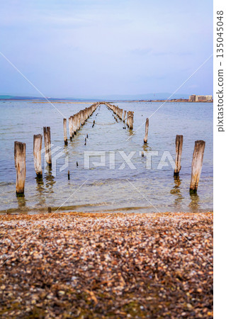 Old wooden pier remains stretch into shallow sea near Pomorie, Bulgaria, with soft blue sky 135045048