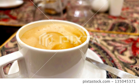 Close-Up of Frothy Coffee in White Cup on Traditional Tablecloth Close-Up of Frothy Coffee in White Cup on Traditional Tablecloth 135045371