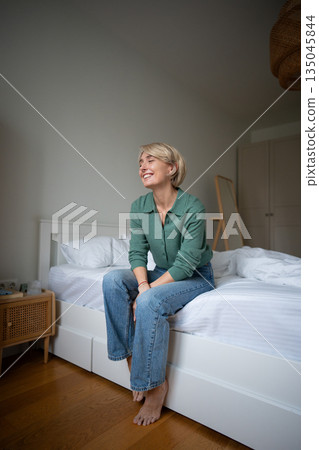 Carefree positive woman sitting on bed in bedroom and smiling brightly, showing self love and peace. 135045844