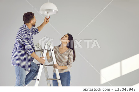 Young man standing on the ladder changing light bulb in the room with his smiling wife. 135045942
