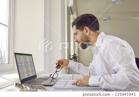 Businessman sitting at table in office looking at report on computer screen 135045943
