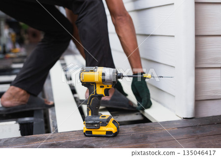 A construction worker is using a cordless power drill to install exterior siding on a house. A construction worker is using a cordless power drill to install exterior siding on a house. 135046417