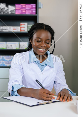 Smiling african american young doctor sitting at the table and making notes 135046708