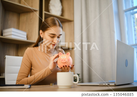 Woman counting savings with piggy bank in home office during daytime Woman counting savings with piggy bank in home office during daytime 135046885