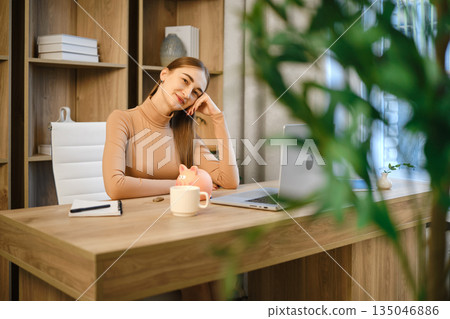 Woman sitting at desk with laptop, mug, and piggy bank in a modern office space Woman sitting at desk with laptop, mug, and piggy bank in a modern office space 135046886