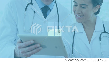Reviewing tablet computer, two doctors in lab coats using stethoscopes analyzing data in exam room Reviewing tablet computer, two doctors in lab coats using stethoscopes analyzing data in exam room 135047652