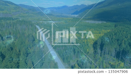 Snaking paved highway winding through conifer forest in mountain valley, with two cars and peaks 135047906