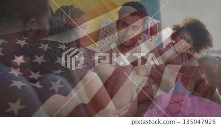 Reclining friends chatting on shore over colorful towels, with pillows, drink cup, US flag overlay 135047928