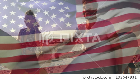 Walking group of friends carrying surfboards on sandy beach, with rocks and American flag overlay 135047931