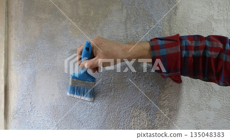 Hand of a man in a plaid shirt holds a blue brush and applies liquid primer on a textured plastered gray wall, close-up, priming a home wall with uneven textured plaster using a brush Hand of a man in a plaid shirt holds a blue brush and applies liquid primer on a textured plastered gray wall, close-up, priming a home wall with uneven textured plaster using a brush 135048383
