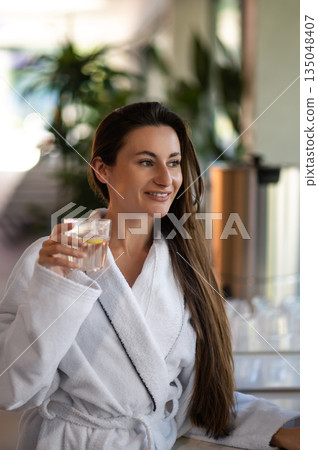 Woman in white bathrobe drinking hot water with lemon 135048407