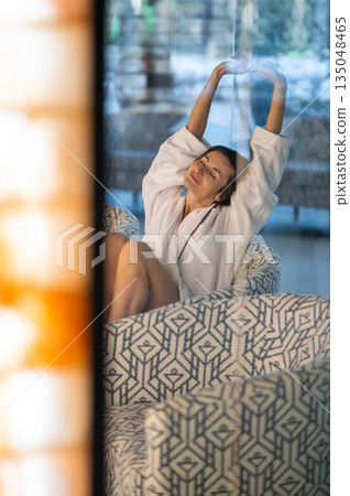 Dark-haired caucasian young woman in a bath robe sitting n a chair Dark-haired caucasian young woman in a bath robe sitting n a chair 135048465