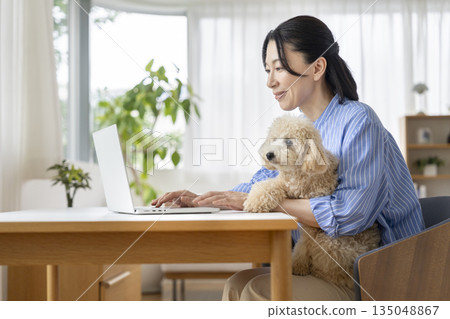 A middle-aged woman working on a computer while holding her pet dog. Image of working from home. 135048867