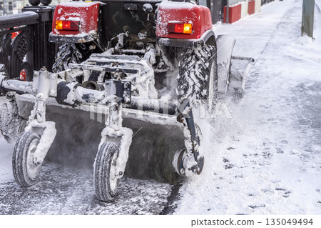 Snowy winter in city. Snow removal machine with rotating brush clears icy sidewalk after heavy snowfall. Concept of climate resilience, public safety, urban winter infrastructure 135049494