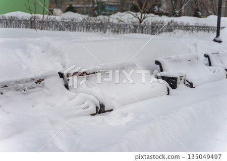 Snowy winter street. Snow covered park benches surrounded by deep snowdrifts during heavy snowfall. Concept of winter abandonment, seasonal challenges, frozen cityscape 135049497