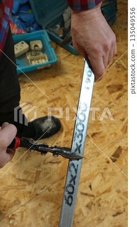 A vertical photo of a worker using metal shears and a narrow silver profile while cutting metal to the required size in a room with OSB flooring, with a toolbox in the background. A vertical photo of a worker using metal shears and a narrow silver profile while cutting metal to the required size in a room with OSB flooring, with a toolbox in the background. 135049556