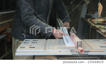 An impersonal carpenter works on a circular saw, cutting a narrow wooden plank lengthwise against the background of an equipped workshop, machine processing and preparation of panels for construction An impersonal carpenter works on a circular saw, cutting a narrow wooden plank lengthwise against the background of an equipped workshop, machine processing and preparation of panels for construction 135049619