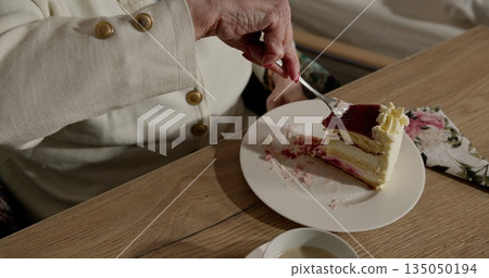 Older woman savoring delicious layered cake. Close-up shot of hand holding fork. Creamy dessert with berry topping on plate. Cozy and relaxed atmosphere. Older woman savoring delicious layered cake. Close-up shot of hand holding fork. Creamy dessert with berry topping on plate. Cozy and relaxed atmosphere. 135050194