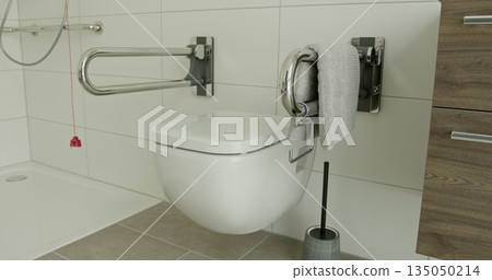 Modern bathroom featuring wall-mounted toilet, safety support bars, and towel. Accessible and functional design promoting independence and safety Modern bathroom featuring wall-mounted toilet, safety support bars, and towel. Accessible and functional design promoting independence and safety 135050214