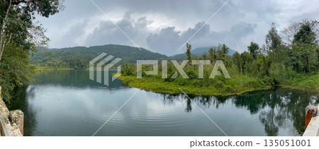 Panoramic view of a tropical mountain lake surrounded by lush green forest and hills, calm water reflections under cloudy sky, peaceful nature landscape 135051001