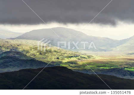 This is a picturesque view in Killarney National Park in Ireland. Sunlight breaks through the dark, stormy sky, illuminating the rolling green hills and valley below to create a breathtaking scene. This is a picturesque view in Killarney National Park in Ireland. Sunlight breaks through the dark, stormy sky, illuminating the rolling green hills and valley below to create a breathtaking scene. 135051476