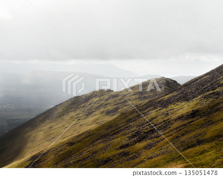 A steep mountain slope covered in grass and rocks ascends into a white sky. 135051478