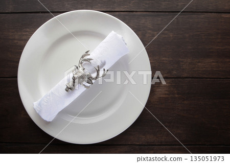 Napkin with serving ring in the form of deer head on white plate. Festive table setting. Top view. Concept for New Year or Christmas. Copy space. Flat lay. Wooden background. 135051973