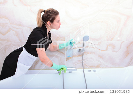 Maid in a uniform cleans the bathroom and the shower faucet. Cleaning services in the house. Hands in protective gloves. The concept of cleanliness and order. 135051985