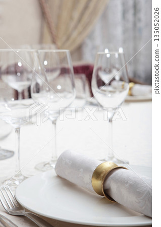 Festive table setting. The napkin is on the plate. A napkin ring. Wine glasses on the table. White plates. White tablecloth with textured pattern. Part of the interior. Festive table setting. The napkin is on the plate. A napkin ring. Wine glasses on the table. White plates. White tablecloth with textured pattern. Part of the interior. 135052026