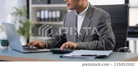 Man calculating finances at home office desk with calculator and papers. Man calculating finances at home office desk with calculator and papers. 135052082