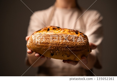 Female baker holding freshly baked artisan bread with seeds, showcasing handmade sourdough loaf in bakery environment with warm light Female baker holding freshly baked artisan bread with seeds, showcasing handmade sourdough loaf in bakery environment with warm light 135052161