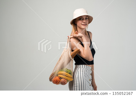 Stylish woman carrying reusable net shopping bag with groceries, eco friendly lifestyle and zero waste concept isolated on light background 135052167