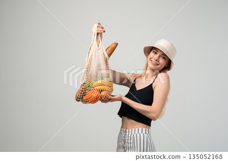 Young woman holding eco friendly net bag with vegetables and bread, sustainable grocery shopping and modern lifestyle concept Young woman holding eco friendly net bag with vegetables and bread, sustainable grocery shopping and modern lifestyle concept 135052168