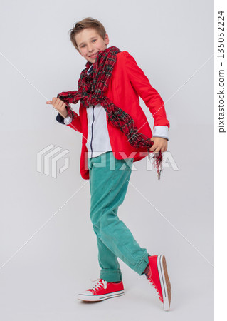 Funny boy in a red jacket, green jeans and sneakers in full growth on a gray background. He waves a long scarf. Funny boy in a red jacket, green jeans and sneakers in full growth on a gray background. He waves a long scarf. 135052224