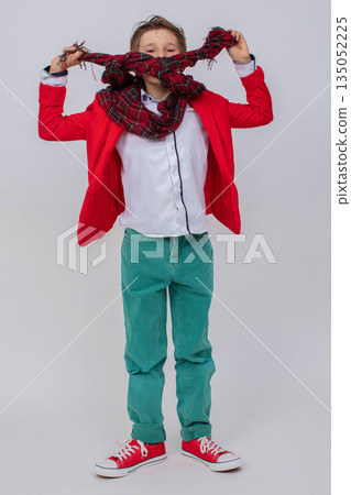 Funny boy in a red jacket, green jeans and sneakers in full growth on a gray background. He hid his face behind a long scarf. Funny boy in a red jacket, green jeans and sneakers in full growth on a gray background. He hid his face behind a long scarf. 135052225