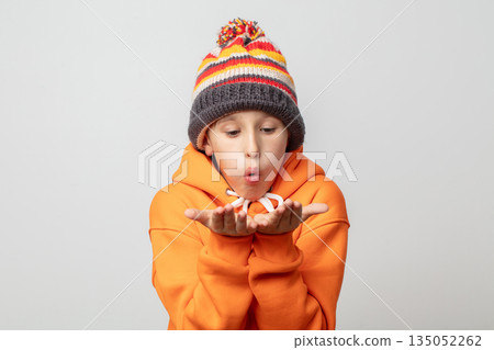 A boy in an orange hoodie and a striped hat blows on his hands. A boy in an orange hoodie and a striped hat blows on his hands. 135052262