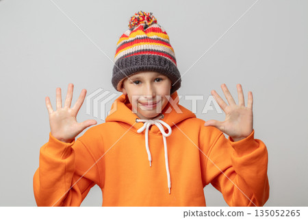 A boy in an orange hoodie and striped hat raised both hands up and showed five fingers. 135052265