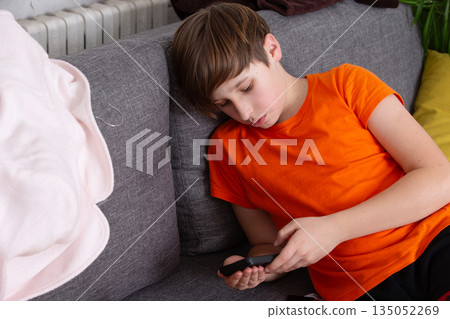 Boy in orange shirt relaxing on gray couch while using smartphone in a cozy living room 135052269