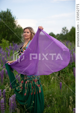 Woman dancing in a lush field wearing a colorful outfit and holding a purple scarf during sunset 135052771