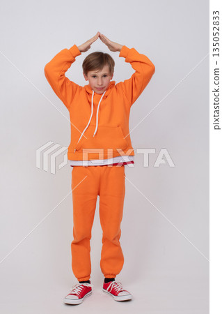 A boy in an orange sweatshirt and orange pants holds his hands in a roof-over-head pose. A boy in an orange sweatshirt and orange pants holds his hands in a roof-over-head pose. 135052833